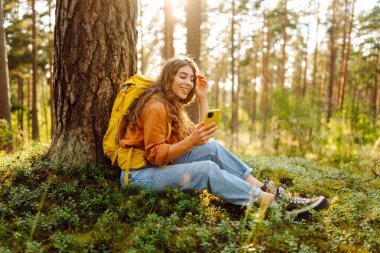Renkli bir sırt çantası ve elinde bir telefonla gezgin bir kadın gün batımında ormanda bir ağacın yanında dinleniyor. Açık hava macerasından zevk alan güzel bir kadın. Seyahat kavramı, blog yazma.