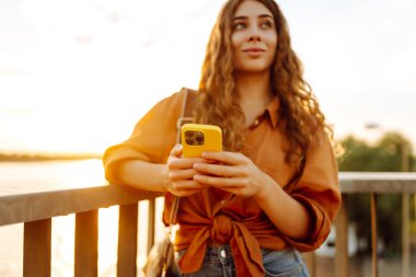 Telefonu olan güzel bir kadın gün batımı seti boyunca yürüyor. Genç bir turist havayı ve blogları sever. Boş zaman konsepti, blog. Etkin yaşam biçimi.