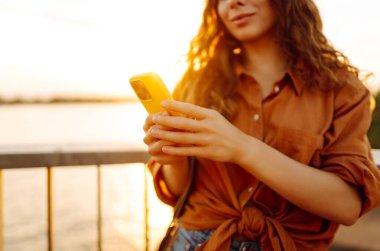 Telefonu olan güzel bir kadın gün batımı seti boyunca yürüyor. Genç bir turist havayı ve blogları sever. Boş zaman konsepti, blog. Etkin yaşam biçimi.