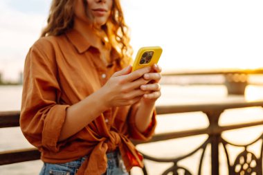 Telefonu olan güzel bir kadın gün batımı seti boyunca yürüyor. Genç bir turist havayı ve blogları sever. Boş zaman konsepti, blog. Etkin yaşam biçimi.