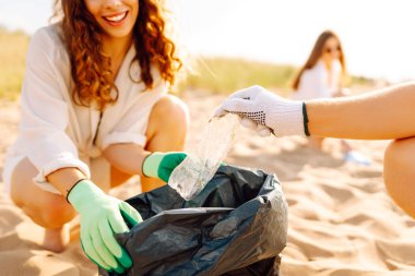 Bir grup genç gönüllü güneşli bir sahilde plastik atık topluyor. Arkadaşları sahildeki çöpleri temizliyor, takım çalışması yapıyorlar. Çevreye yardım etmek. Gönüllü olma kavramı.