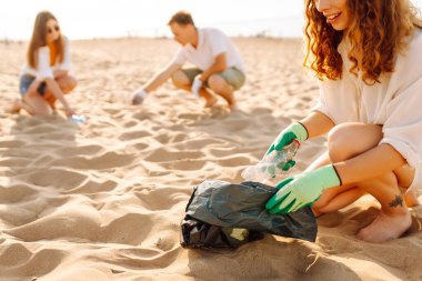 Bir grup genç gönüllü güneşli bir sahilde plastik atık topluyor. Arkadaşları sahildeki çöpleri temizliyor, takım çalışması yapıyorlar. Çevreye yardım etmek. Gönüllü olma kavramı.