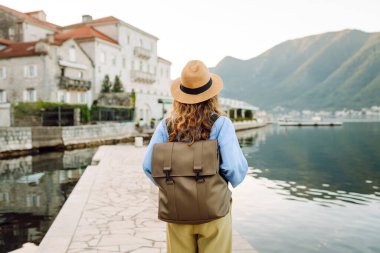 Şapkalı ve sırt çantalı genç bir turistin duvar boyunca yürüdüğü arka plan görüntüsü. Güzel bir kadın deniz manzarasını ve dağları sever. Turizm, keyif ve rahatlama kavramı.