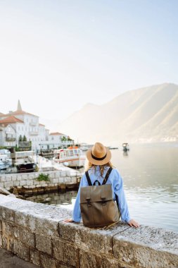 Şapkalı ve sırt çantalı genç bir turistin duvar boyunca yürüdüğü arka plan görüntüsü. Güzel bir kadın deniz manzarasını ve dağları sever. Turizm, keyif ve rahatlama kavramı.