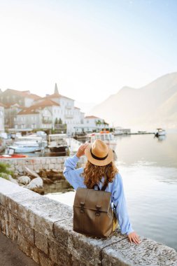 Şapkalı ve sırt çantalı genç bir turistin duvar boyunca yürüdüğü arka plan görüntüsü. Güzel bir kadın deniz manzarasını ve dağları sever. Turizm, keyif ve rahatlama kavramı.