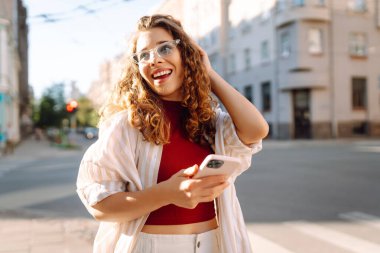 Güneşli bir caddede telefonu olan neşeli bir kadın. Güzel turist sohbetleri, dışarıda blog yazışmaları. Moda konsepti. Yaşam biçimi.