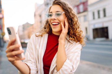 Güneşli bir caddede telefonu olan neşeli bir kadın. Güzel turist sohbetleri, dışarıda blog yazışmaları. Moda konsepti. Yaşam biçimi.