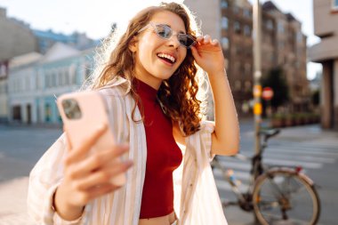 Güneşli bir caddede telefonu olan neşeli bir kadın. Güzel turist sohbetleri, dışarıda blog yazışmaları. Moda konsepti. Yaşam biçimi.
