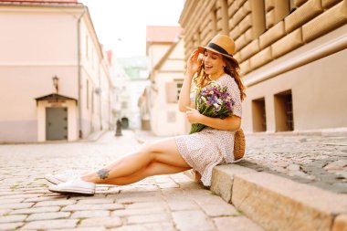 Şapkalı mutlu bir kadın ve güzel bir buket kır çiçeğiyle küçük bir kasabanın güneşli sokaklarının tadını çıkarıyor. Genç bayan turist dışarı çıkıp eğleniyor. Rahatlama, eğlence, güzellik kavramı..