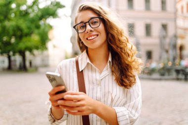 Şık giyinmiş, elinde telefonla güneşli şehir sokaklarında yürüyen genç bir kadının portresi. Telefon mesajı atan, blog yazan güzel bir turist. Hafta sonu konsepti, iletişim. Yaşam biçimi.