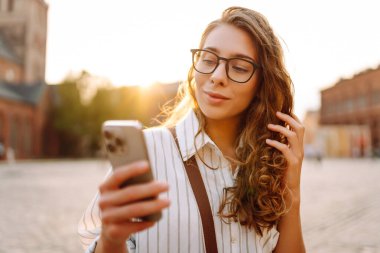Şık giyinmiş, elinde telefonla güneşli şehir sokaklarında yürüyen genç bir kadının portresi. Telefon mesajı atan, blog yazan güzel bir turist. Hafta sonu konsepti, iletişim. Yaşam biçimi.