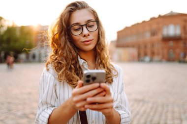 Şık giyinmiş, elinde telefonla güneşli şehir sokaklarında yürüyen genç bir kadının portresi. Telefon mesajı atan, blog yazan güzel bir turist. Hafta sonu konsepti, iletişim. Yaşam biçimi.