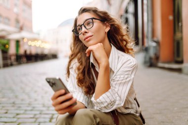 Şık giyinmiş, elinde telefonla güneşli şehir sokaklarında yürüyen genç bir kadının portresi. Telefon mesajı atan, blog yazan güzel bir turist. Hafta sonu konsepti, iletişim. Yaşam biçimi.