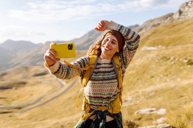 Sarı sırt çantalı genç bir kadın elinde telefonla güneşli bir yürüyüş gününün tadını çıkarıyor. Güzel bir kadın selfie çekiyor ve çarpıcı dağ manzarasının tadını çıkarıyor. Macera ve günlük kavramı.