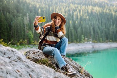 Elinde telefon olan güzel bir kadın bir kayanın üzerindeki dağ manzarasının tadını çıkarıyor. Bir gezgin blogu, doğada selfie çeker. Seyahat konsepti, keyif, blog yazma.