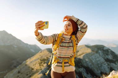 Yürüyüş ekipmanlarıyla dağda selfie çeken bir yürüyüşçü. Neşeli kadın özgürlüğü hissediyor ve gün batımının tadını çıkarıyor. Yürüyüş, macera konsepti. Etkin yaşam biçimi.