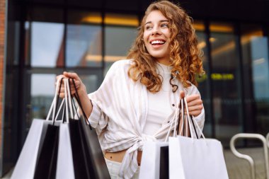 Alışveriş yaptıktan sonra çantalarını tutan modern giysili mutlu kadın portresi. Genç bir kadın dışarıda eğleniyor ve yeni alışverişlerden zevk alıyor. Tüketim kavramı, alışveriş.