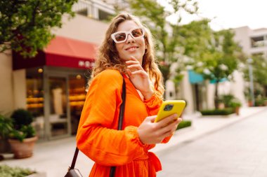 Şık, parlak elbiseli güzel bir kadın telefonuyla güneşli bir set boyunca yürüyor. Genç bir turist blogu, tatilde havanın tadını çıkarıyor. Seyahat kavramı, blog yazma.