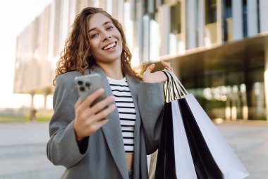 İnternetten alışveriş! Alışveriş merkezinin yakınında telefonu olan mutlu bir kadın. Alışverişten sonra akıllı telefonlu genç bir kadın, açık havada günbatımının tadını çıkarıyor. Tüketim kavramı.
