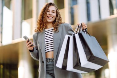 İnternetten alışveriş! Alışveriş merkezinin yakınında telefonu olan mutlu bir kadın. Alışverişten sonra akıllı telefonlu genç bir kadın, açık havada günbatımının tadını çıkarıyor. Tüketim kavramı.