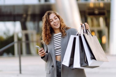 İnternetten alışveriş! Alışveriş merkezinin yakınında telefonu olan mutlu bir kadın. Alışverişten sonra akıllı telefonlu genç bir kadın, açık havada günbatımının tadını çıkarıyor. Tüketim kavramı.