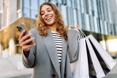 İnternetten alışveriş! Alışveriş merkezinin yakınında telefonu olan mutlu bir kadın. Alışverişten sonra akıllı telefonlu genç bir kadın, açık havada günbatımının tadını çıkarıyor. Tüketim kavramı.