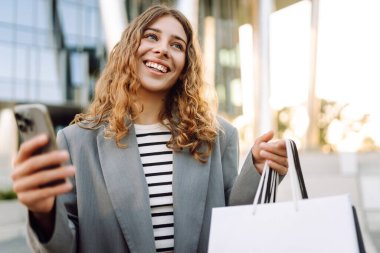İnternetten alışveriş! Alışveriş merkezinin yakınında telefonu olan mutlu bir kadın. Alışverişten sonra akıllı telefonlu genç bir kadın, açık havada günbatımının tadını çıkarıyor. Tüketim kavramı.