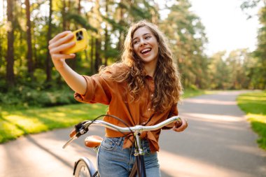 Selfie zamanı! Parkta selfie çeken, telefonla bisiklet süren genç bir kadın. Güneşli bir parkta eğlenen mutlu kadın. Etkinlik, eğlence ve hafta sonu konsepti.