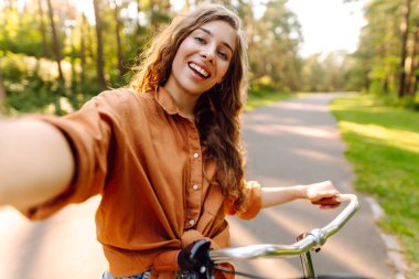 Selfie zamanı! Parkta selfie çeken, telefonla bisiklet süren genç bir kadın. Güneşli bir parkta eğlenen mutlu kadın. Etkinlik, eğlence ve hafta sonu konsepti.
