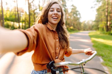 Selfie zamanı! Parkta selfie çeken, telefonla bisiklet süren genç bir kadın. Güneşli bir parkta eğlenen mutlu kadın. Etkinlik, eğlence ve hafta sonu konsepti.