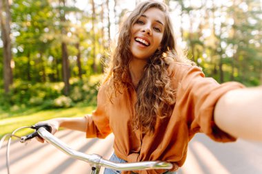 Selfie zamanı! Parkta selfie çeken, telefonla bisiklet süren genç bir kadın. Güneşli bir parkta eğlenen mutlu kadın. Etkinlik, eğlence ve hafta sonu konsepti.