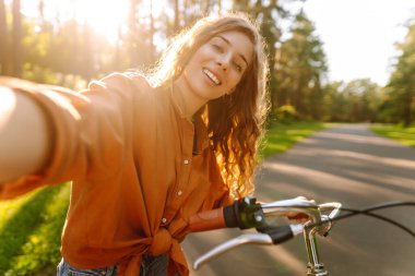 Selfie zamanı! Parkta selfie çeken, telefonla bisiklet süren genç bir kadın. Güneşli bir parkta eğlenen mutlu kadın. Etkinlik, eğlence ve hafta sonu konsepti.