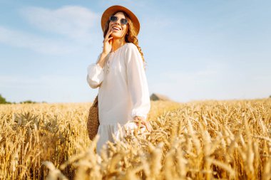 Şapkalı gülümseyen bir kadın altın buğday tarlasında yürüyor. Güzel bir kadın manzaranın tadını çıkarır ve açık havada özgürlüğü hisseder. Yürüme ve eğlenme konsepti.