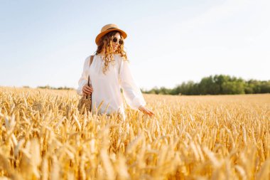 Şapkalı gülümseyen bir kadın altın buğday tarlasında yürüyor. Güzel bir kadın manzaranın tadını çıkarır ve açık havada özgürlüğü hisseder. Yürüme ve eğlenme konsepti.