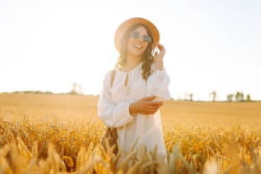 Şapkalı gülümseyen bir kadın altın buğday tarlasında yürüyor. Güzel bir kadın manzaranın tadını çıkarır ve açık havada özgürlüğü hisseder. Yürüme ve eğlenme konsepti.