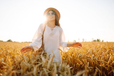 Şapkalı gülümseyen bir kadın altın buğday tarlasında yürüyor. Güzel bir kadın manzaranın tadını çıkarır ve açık havada özgürlüğü hisseder. Yürüme ve eğlenme konsepti.