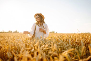 Şapkalı gülümseyen bir kadın altın buğday tarlasında yürüyor. Güzel bir kadın manzaranın tadını çıkarır ve açık havada özgürlüğü hisseder. Yürüme ve eğlenme konsepti.