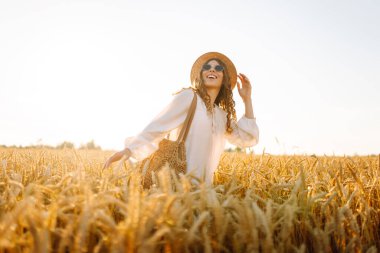 Şapkalı gülümseyen bir kadın altın buğday tarlasında yürüyor. Güzel bir kadın manzaranın tadını çıkarır ve açık havada özgürlüğü hisseder. Yürüme ve eğlenme konsepti.