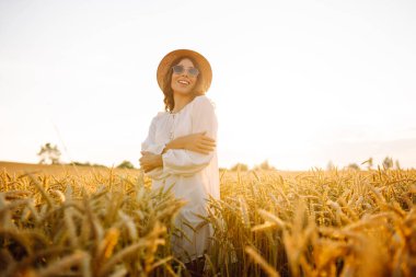 Şapkalı gülümseyen bir kadın altın buğday tarlasında yürüyor. Güzel bir kadın manzaranın tadını çıkarır ve açık havada özgürlüğü hisseder. Yürüme ve eğlenme konsepti.