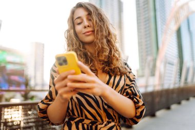 Bir metropolün arka planında telefonu olan şık giyinmiş neşeli bir kadın. Genç bir turist blogu güneşli bir günde yürüyüşe çıkıyor. Turizm, keyif konsepti.