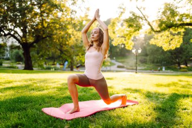 Sunny Park 'ta yoga minderi üzerinde egzersiz yapan genç bir kadın. Spor kıyafetli kadın dışarıda yoga yapıyor. Spor konsepti. Etkin yaşam biçimi.