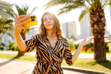 Telefonla konuşan güzel bir kadın, arka planda park halindeyken selfie çekiyor. Telefonla blog yazan genç bir kadın güneşli bir günün tadını çıkarıyor. Turizm, eğlence ve blog konsepti.