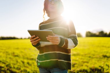 Dijital tabletli genç kadın çiftçi, tarım alanındaki genç bitkileri inceliyor. Kadın tarım uzmanı elinde bir tablet tutuyor ve filizlerin kalitesini kontrol ediyor. Bahçe konsepti.