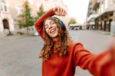 Selfie zamanı! Kablosuz kulaklık takan, müzik dinleyen ve şehir ortamında yürüyen genç bir kadın. Yürüyüşün ve güneşli havanın tadını çıkaran güzel bir turist. Blogculuk, teknoloji, turizm kavramı.