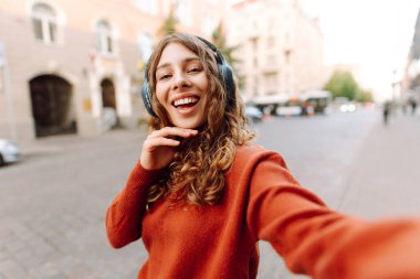Selfie zamanı! Kablosuz kulaklık takan, müzik dinleyen ve şehir ortamında yürüyen genç bir kadın. Yürüyüşün ve güneşli havanın tadını çıkaran güzel bir turist. Blogculuk, teknoloji, turizm kavramı.