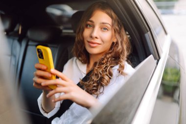 Arabanın arka koltuğunda oturan telefonu olan genç bir iş kadınının portresi. Yolculuk sırasında arabada mesajlaşan ve rahatlatan güzel bir kadın. Ulaşım, eğlence ve teknoloji konsepti.