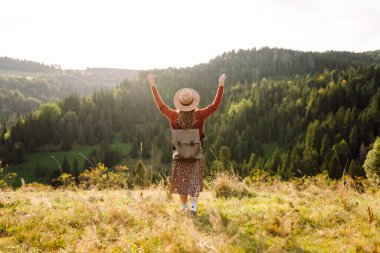 Parlak yeşil vadiyi gezen sırt çantalı güzel bir turist. Şapkalı genç bir kadın günbatımının ve doğanın tadını çıkarıyor. Seyahat, doğa ve özgürlük kavramı. Etkin yaşam biçimi.