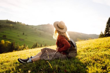 Parlak yeşil vadiyi gezen sırt çantalı güzel bir turist. Şapkalı genç bir kadın günbatımının ve doğanın tadını çıkarıyor. Seyahat, doğa ve özgürlük kavramı. Etkin yaşam biçimi.