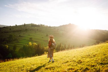 Parlak yeşil vadiyi gezen sırt çantalı güzel bir turist. Şapkalı genç bir kadın günbatımının ve doğanın tadını çıkarıyor. Seyahat, doğa ve özgürlük kavramı. Etkin yaşam biçimi.