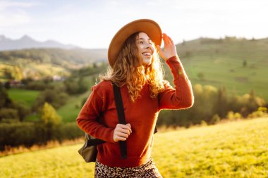 Parlak yeşil vadiyi gezen sırt çantalı güzel bir turist. Şapkalı genç bir kadın günbatımının ve doğanın tadını çıkarıyor. Seyahat, doğa ve özgürlük kavramı. Etkin yaşam biçimi.